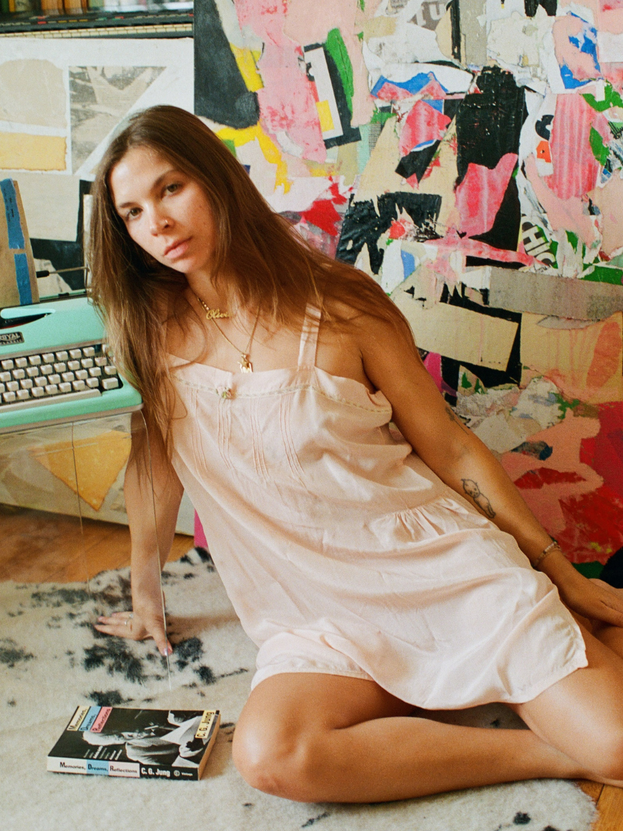 Woman in a white dress sitting on the floor with a colorful abstract painting and typewriter in the background.