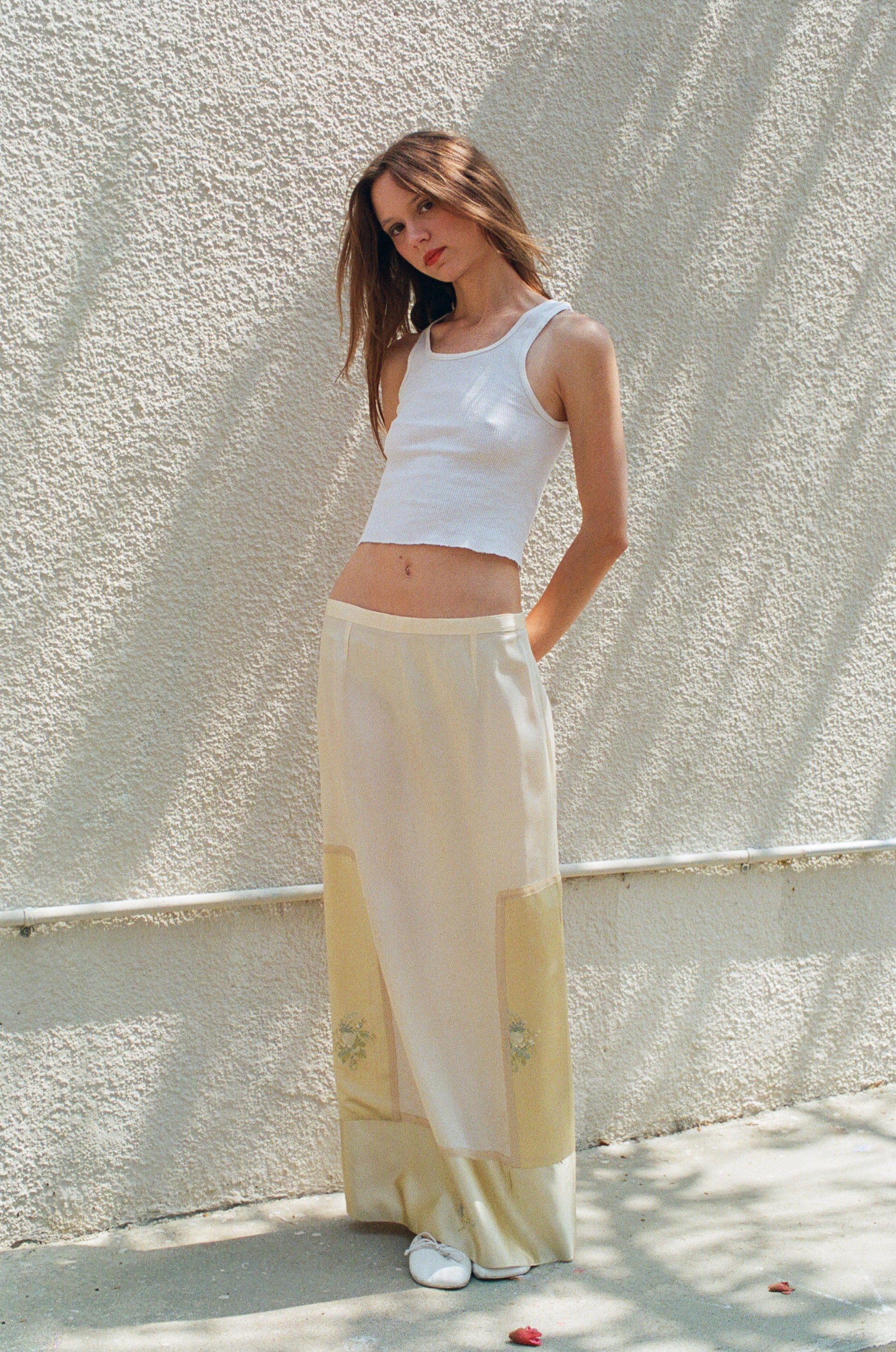 Woman wearing a white sleeveless top and light-colored pants against a textured wall.