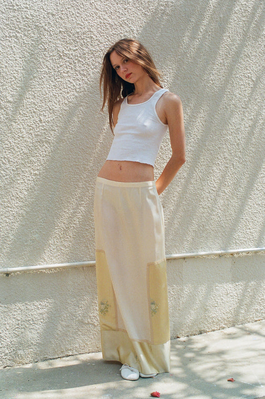 Woman wearing a white sleeveless top and light-colored pants against a textured wall.