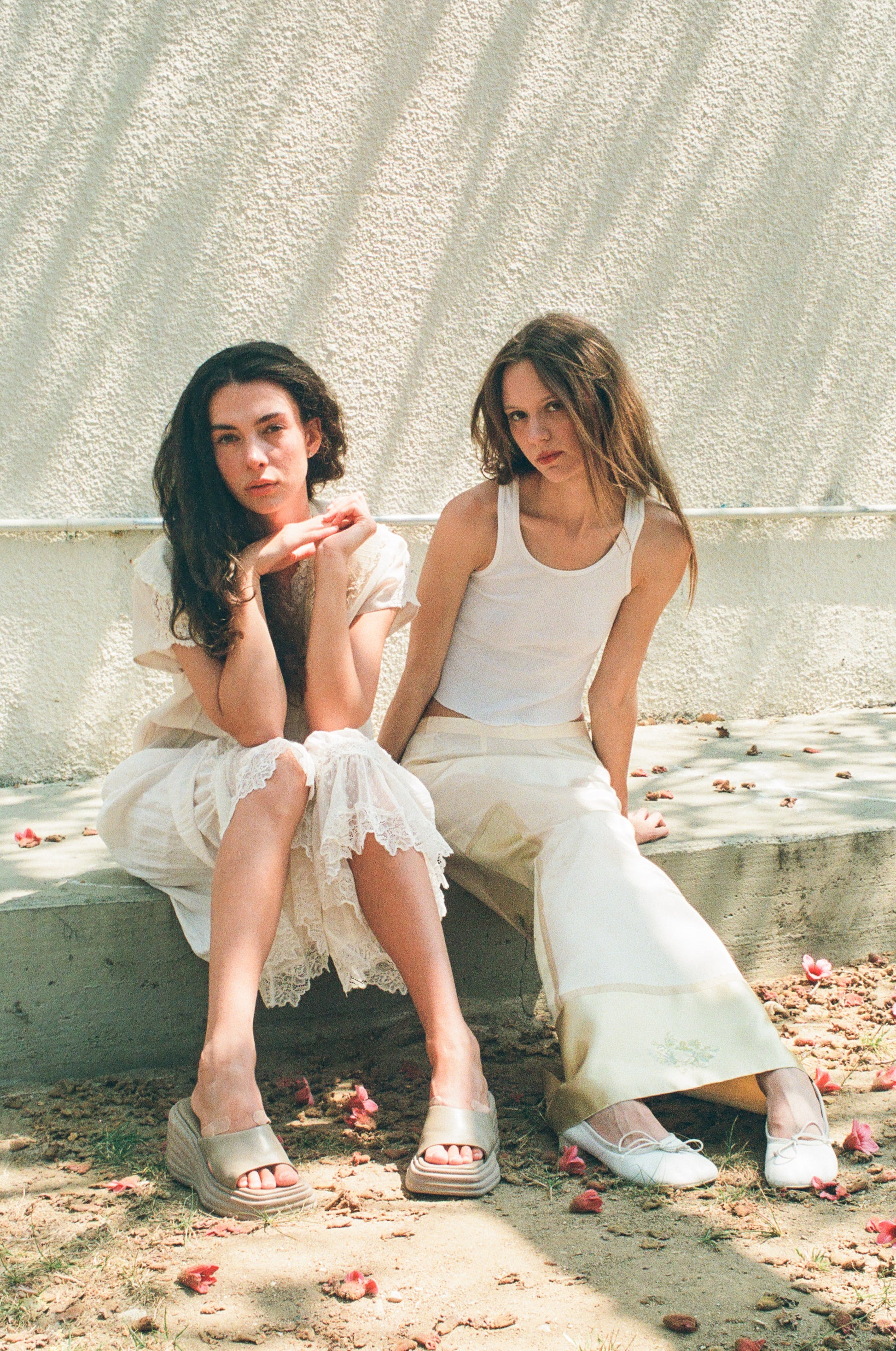 Two women in white dresses sitting on a stone bench with a textured wall in the background.