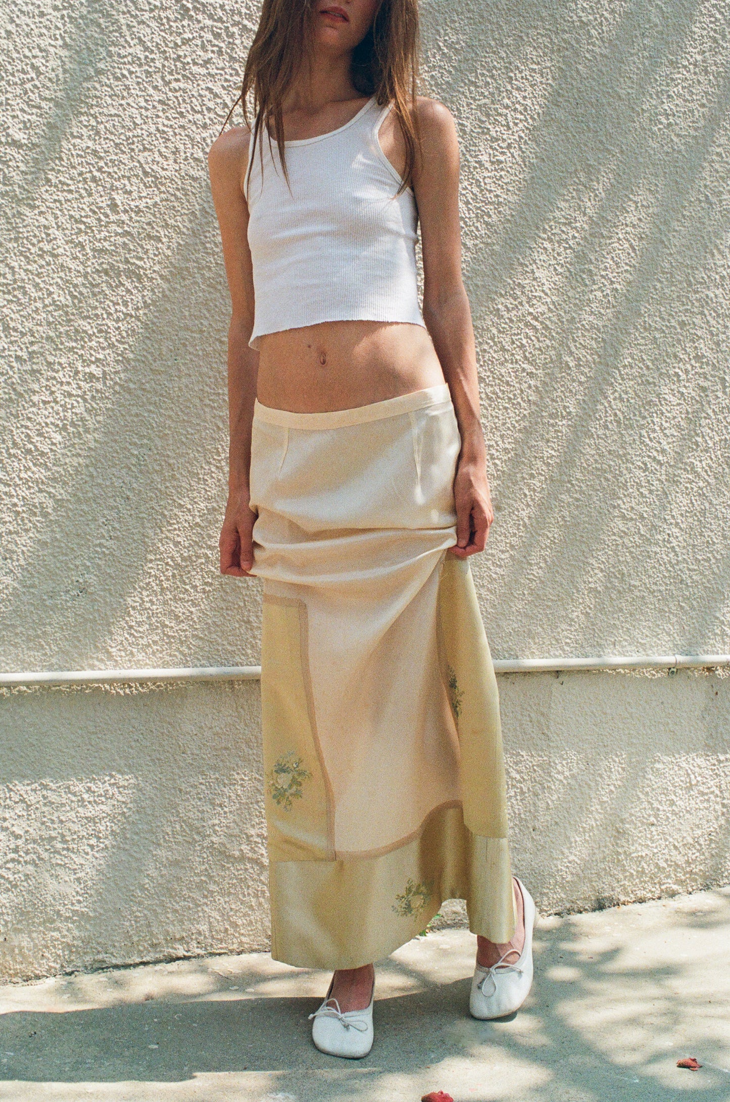 Woman wearing a white tank top and beige skirt against a light wall.