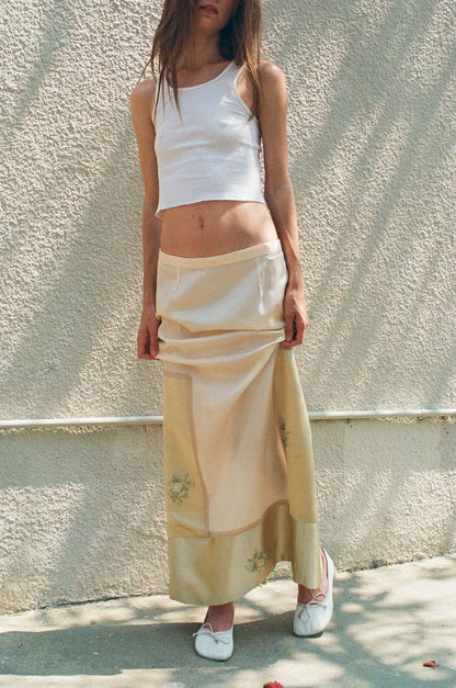 Woman wearing a white tank top and beige skirt against a light wall.