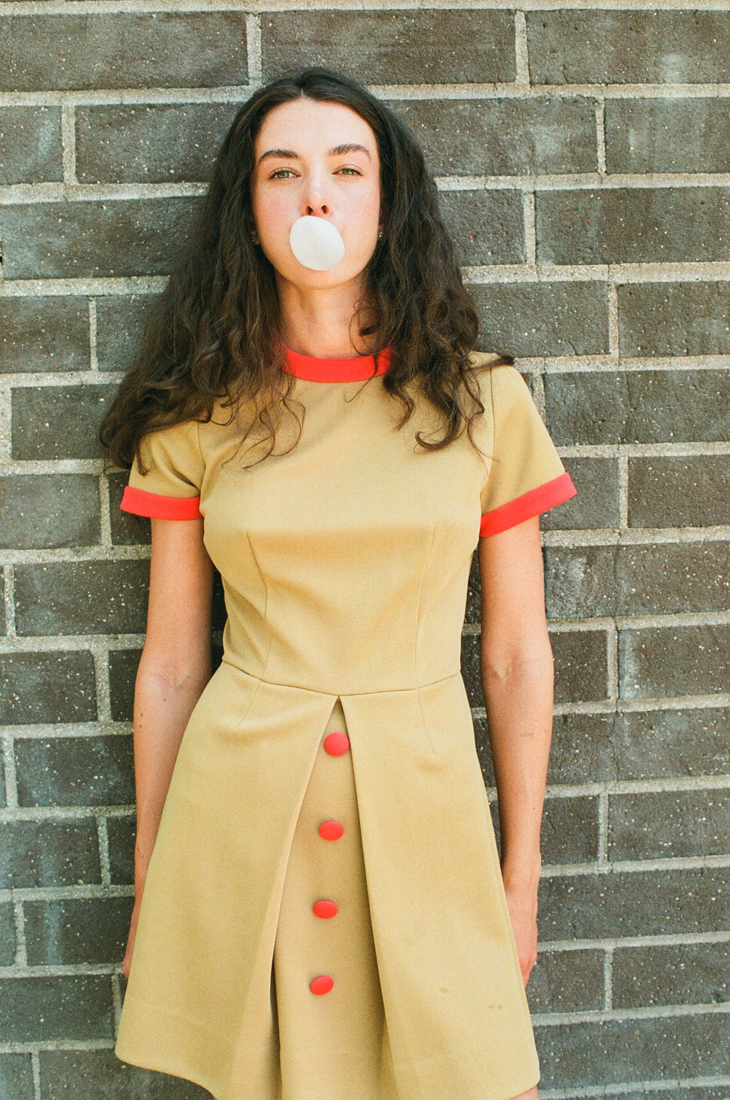 Woman in a beige dress with red accents blowing bubble gum against a brick wall.