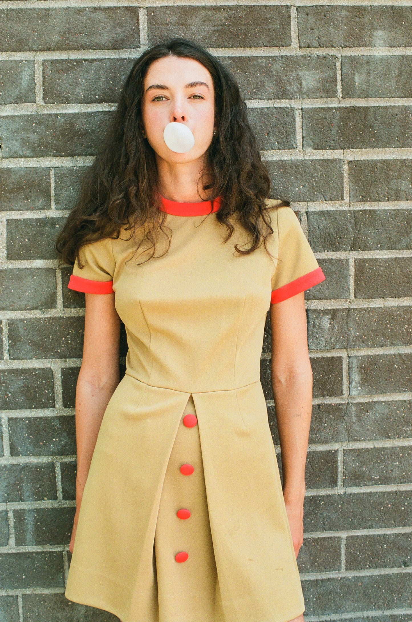 Woman in a beige dress with red accents blowing bubble gum against a brick wall.