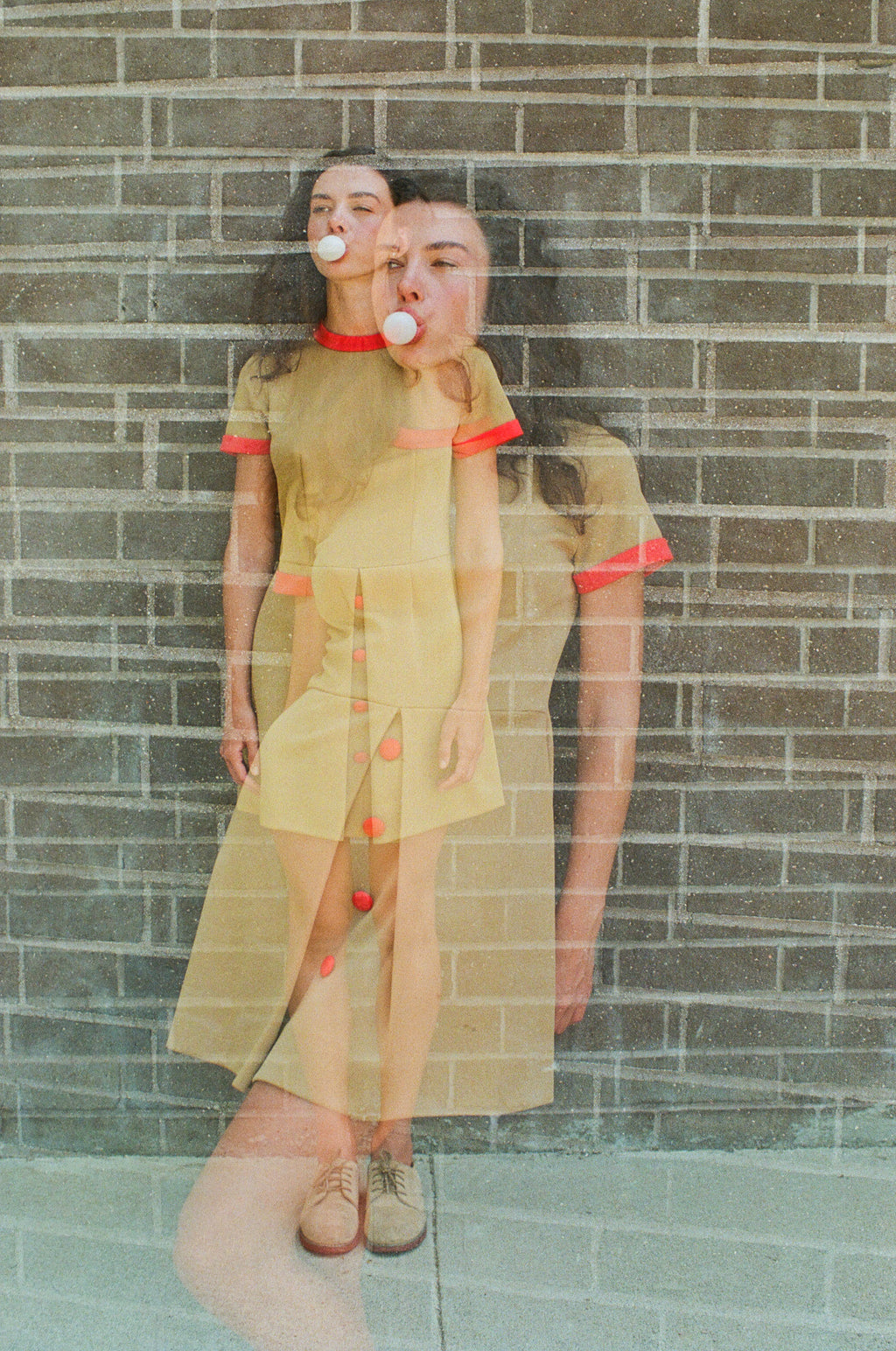 Woman in a yellow dress with red accents standing against a brick wall.