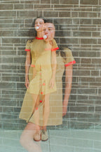 Woman in a yellow dress with red accents standing against a brick wall.