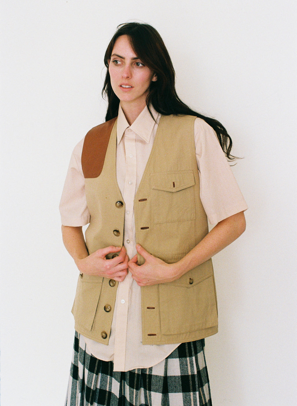 Woman wearing a beige vest over a white shirt with a plaid skirt on a white background