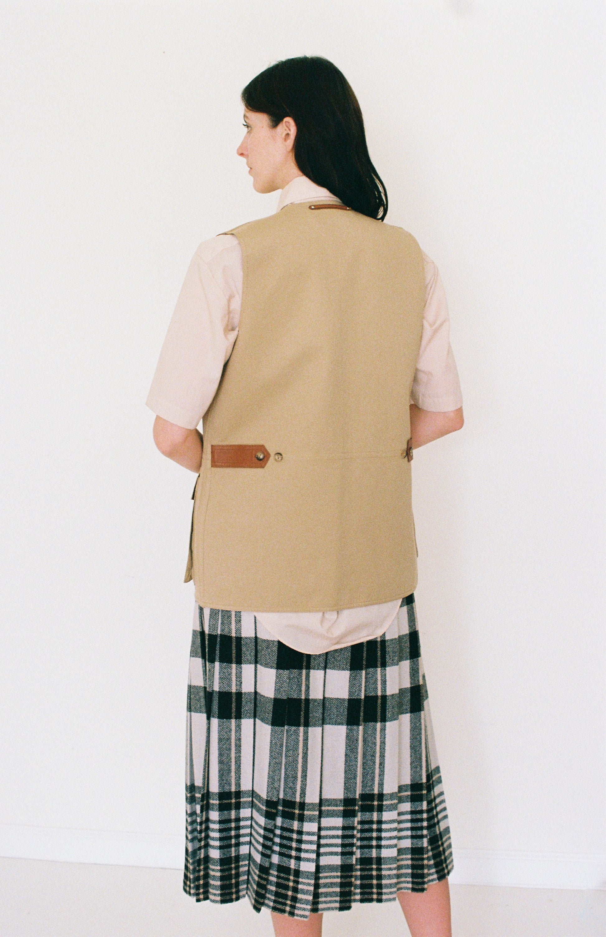 Woman wearing a beige vest over a light shirt and plaid skirt on a white background