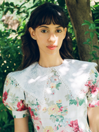 Woman wearing a floral blouse standing outdoors with greenery in the background