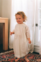 Child in a long, light-colored dress standing in a room with white curtains and a patterned rug.