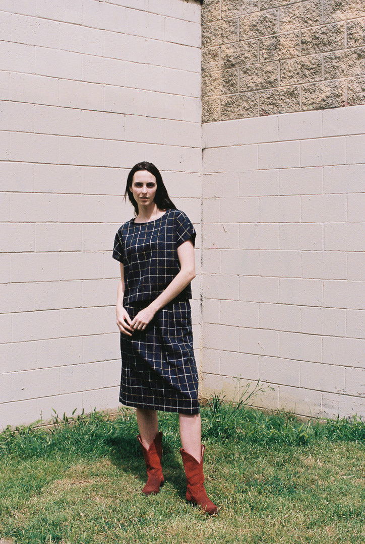 Woman in a black checkered dress and red boots standing against a light-colored wall.