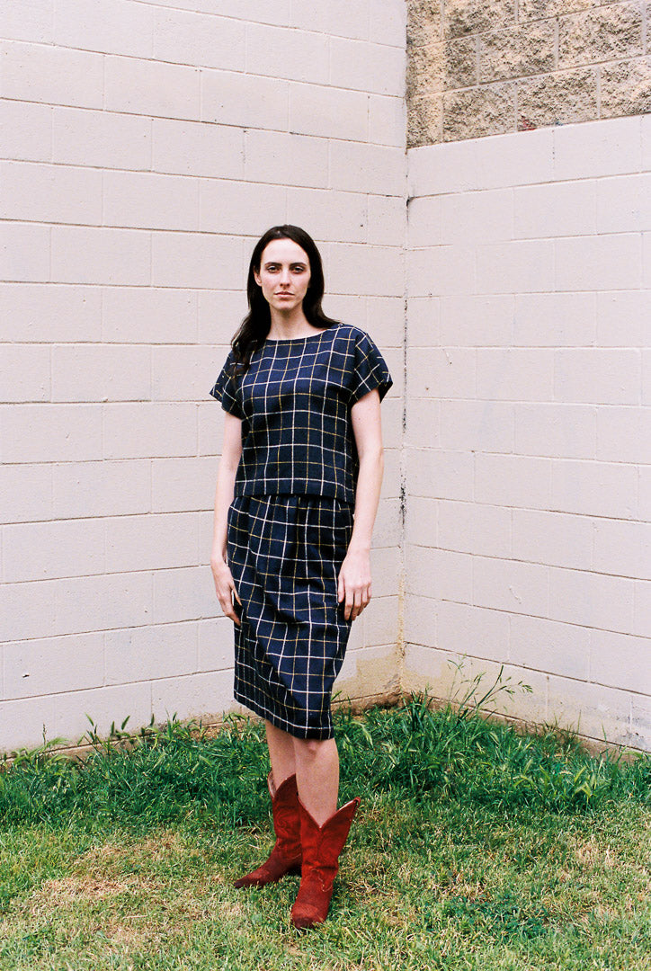 Woman wearing a plaid dress and red boots standing against a white wall.