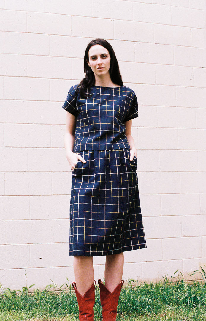 Woman wearing a blue checkered dress with brown boots standing against a white wall.