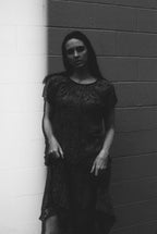 Black and white photo of a person wearing a dark lace dress against a brick wall.
