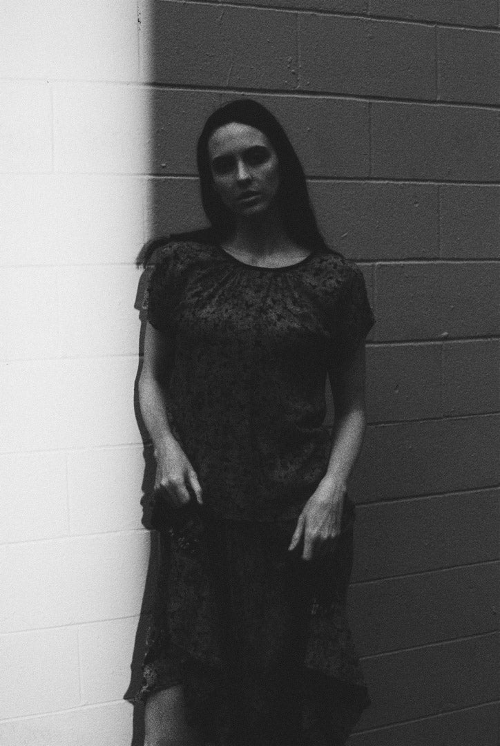 Black and white photo of a person wearing a dark lace dress against a brick wall.