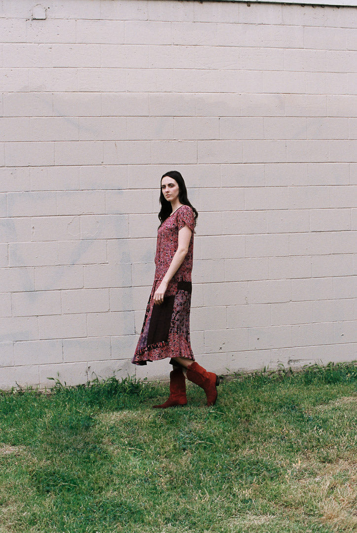 Woman in a patterned dress and red boots standing in front of a light-colored wall.