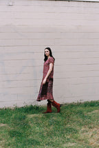 Woman in a patterned dress and red boots standing in front of a light-colored wall.