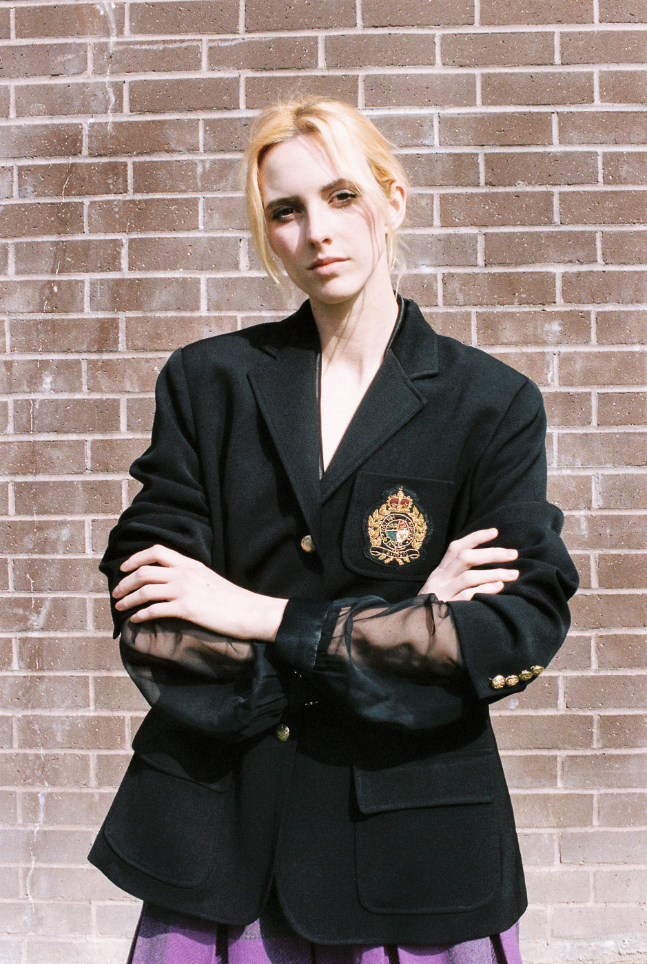 Person wearing a black blazer with a visible emblem against a brick wall