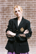 Person wearing a black blazer with a visible emblem against a brick wall