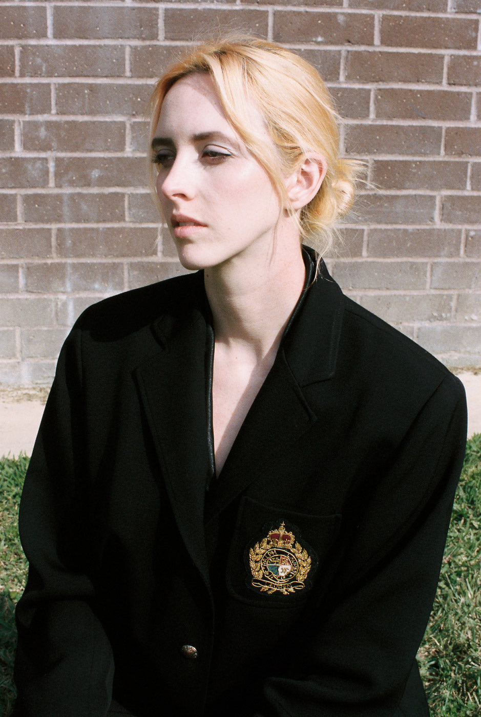 Person wearing a black coat with a visible emblem against a brick wall.