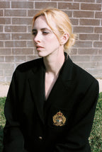 Person wearing a black coat with a visible emblem against a brick wall.