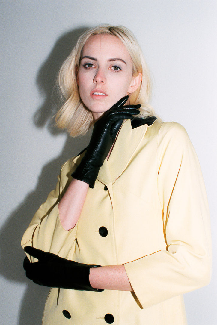 Woman wearing a yellow coat with black buttons and black gloves against a plain background