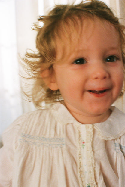 Child with blonde hair wearing a light-colored dress against a neutral background