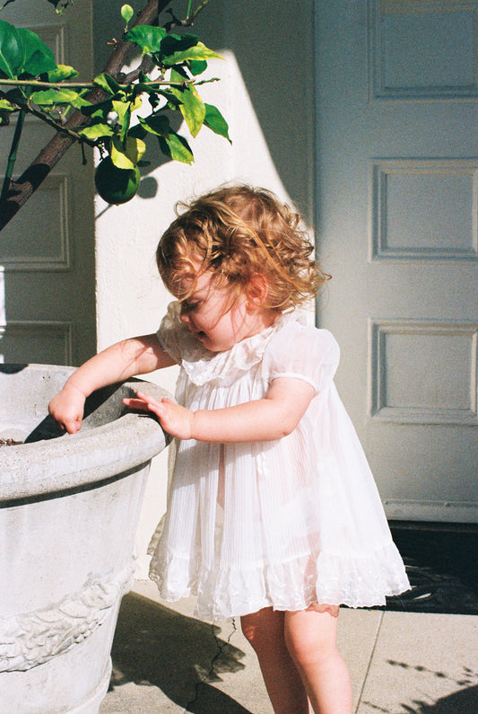 Child in a white dress standing next to a decorative stone planter outdoors.