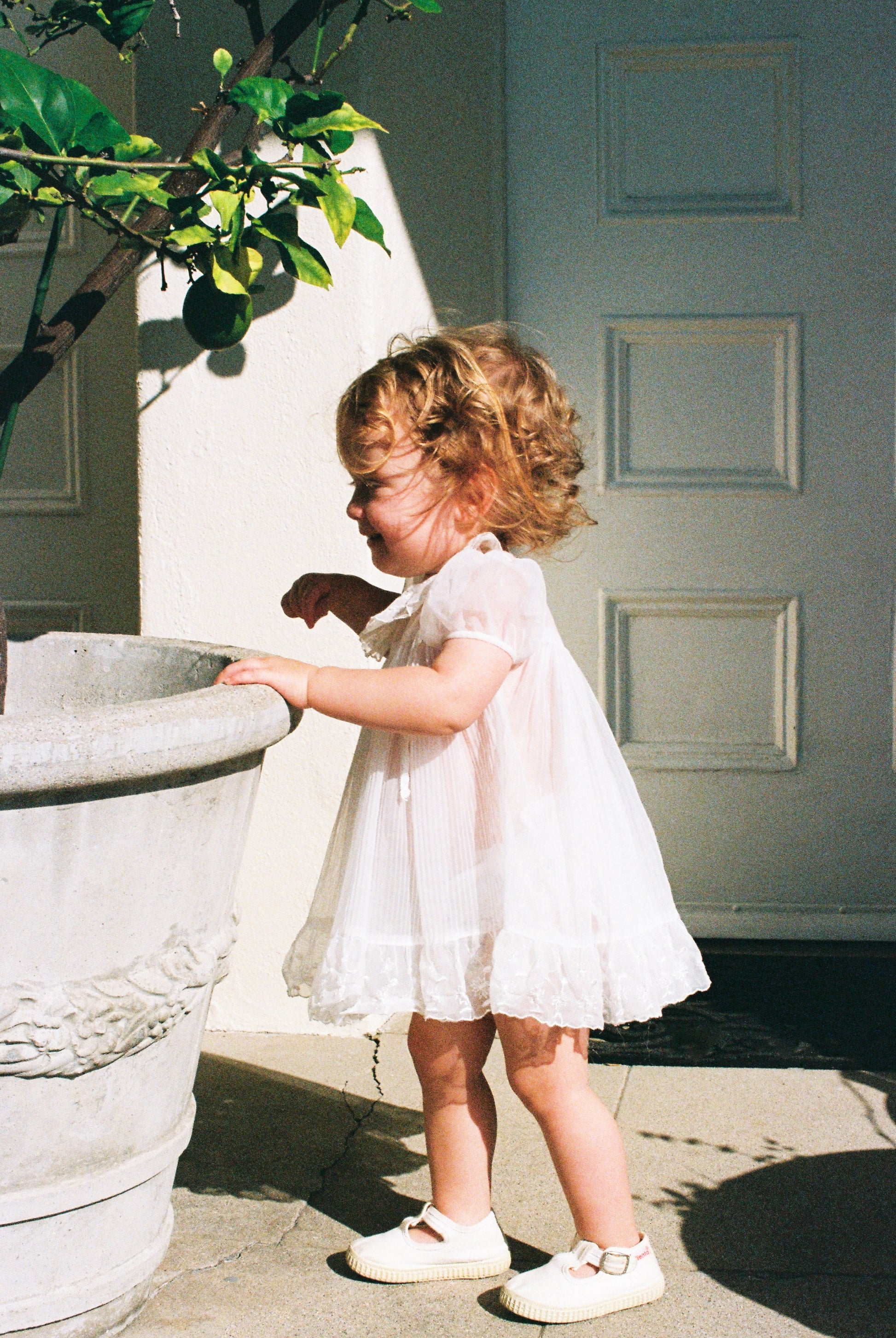 Young girl in a white dress standing next to a stone planter outdoors.