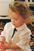 Child wearing a vintage white dress with a black bow in a home setting