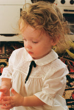 Child wearing a vintage white dress with a black bow in a home setting