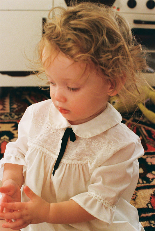 Child wearing a vintage white dress with a black bow in a home setting