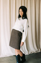 Woman wearing a white blouse, brown skirt, and black boots against a plain background