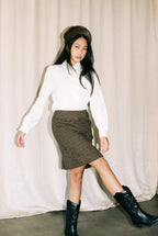Woman wearing a white shirt, brown skirt, and black boots against a plain background