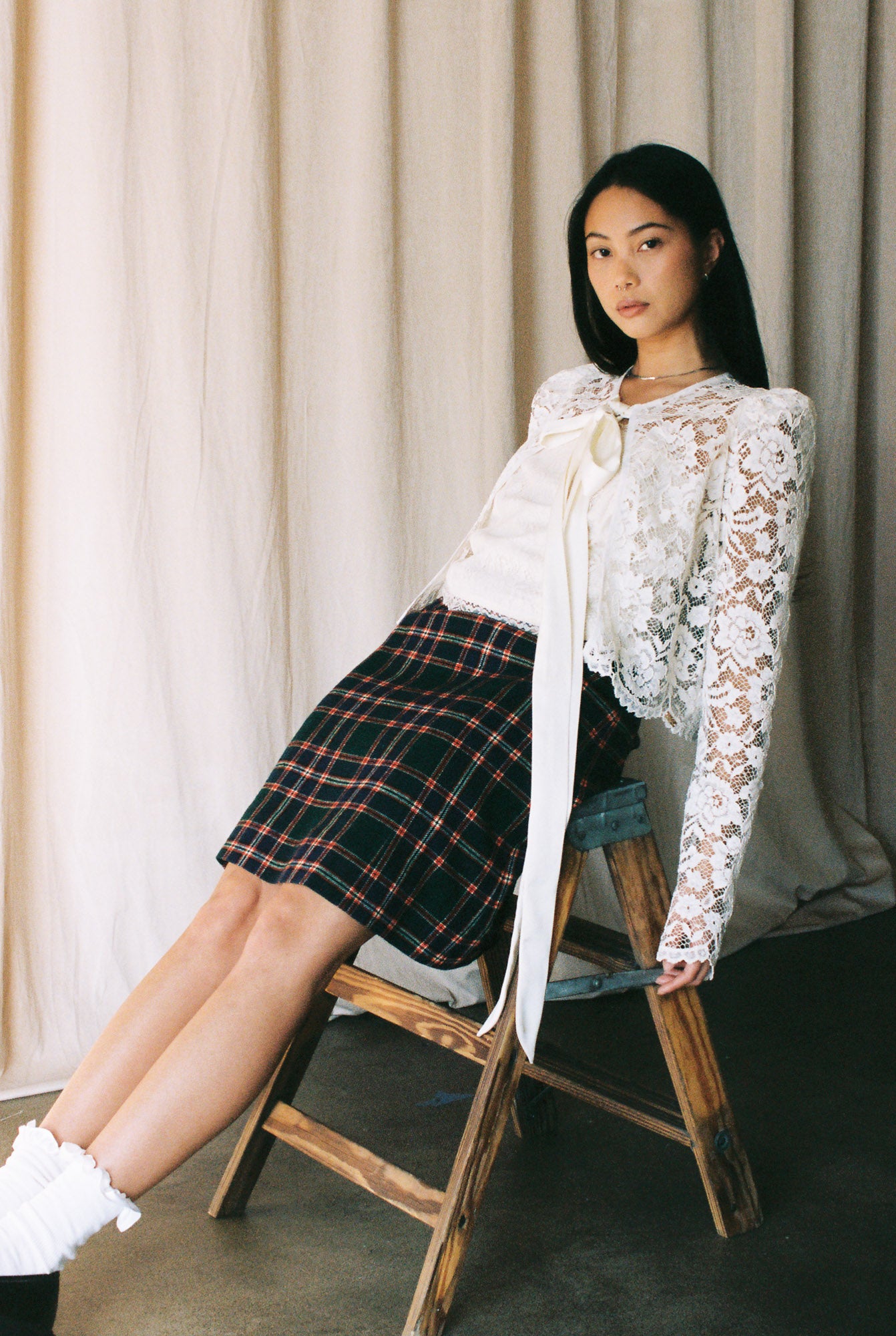 Woman sitting on a wooden chair wearing a white lace top and plaid skirt against a neutral curtain background.
