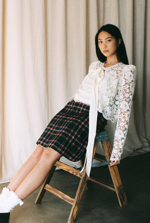 Woman sitting on a wooden chair wearing a white lace top and plaid skirt against a neutral curtain background.