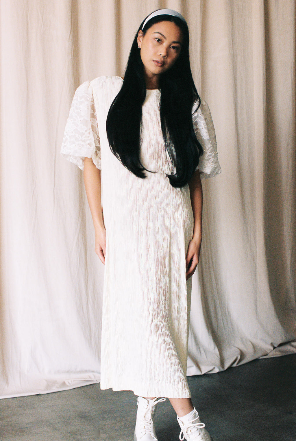 Woman wearing a white dress with lace sleeves against a beige curtain background