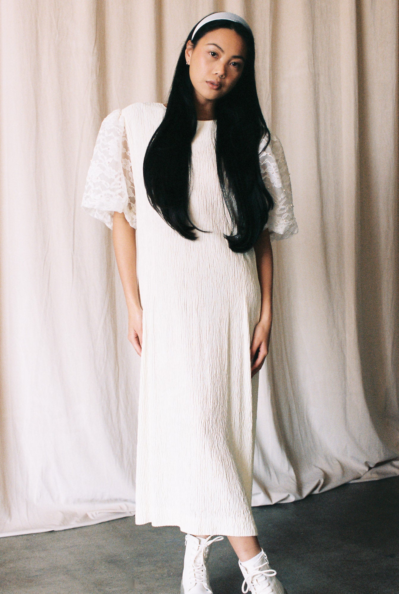 Woman wearing a white dress with lace sleeves against a beige curtain background