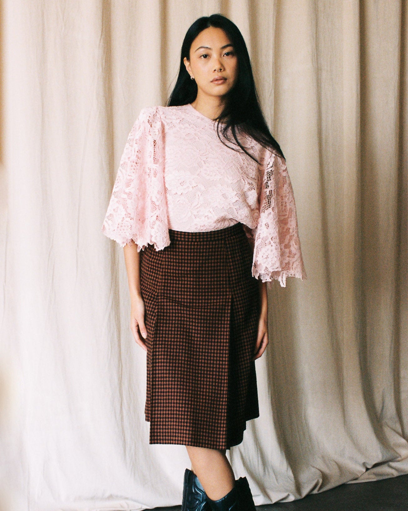 Woman wearing a pink lace top and brown plaid skirt with black boots against a beige curtain background