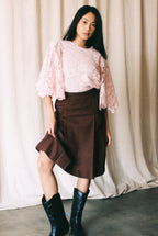 Woman wearing a pink lace top and brown checkered skirt with black boots against a beige curtain background