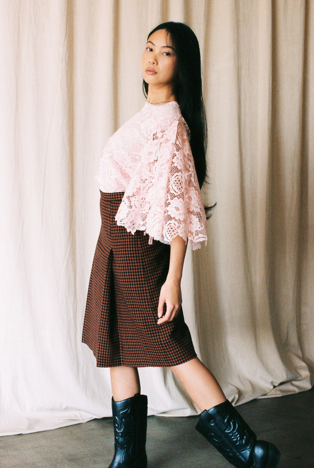 Woman wearing a pink lace top, brown checkered skirt, and black boots against a beige curtain background.
