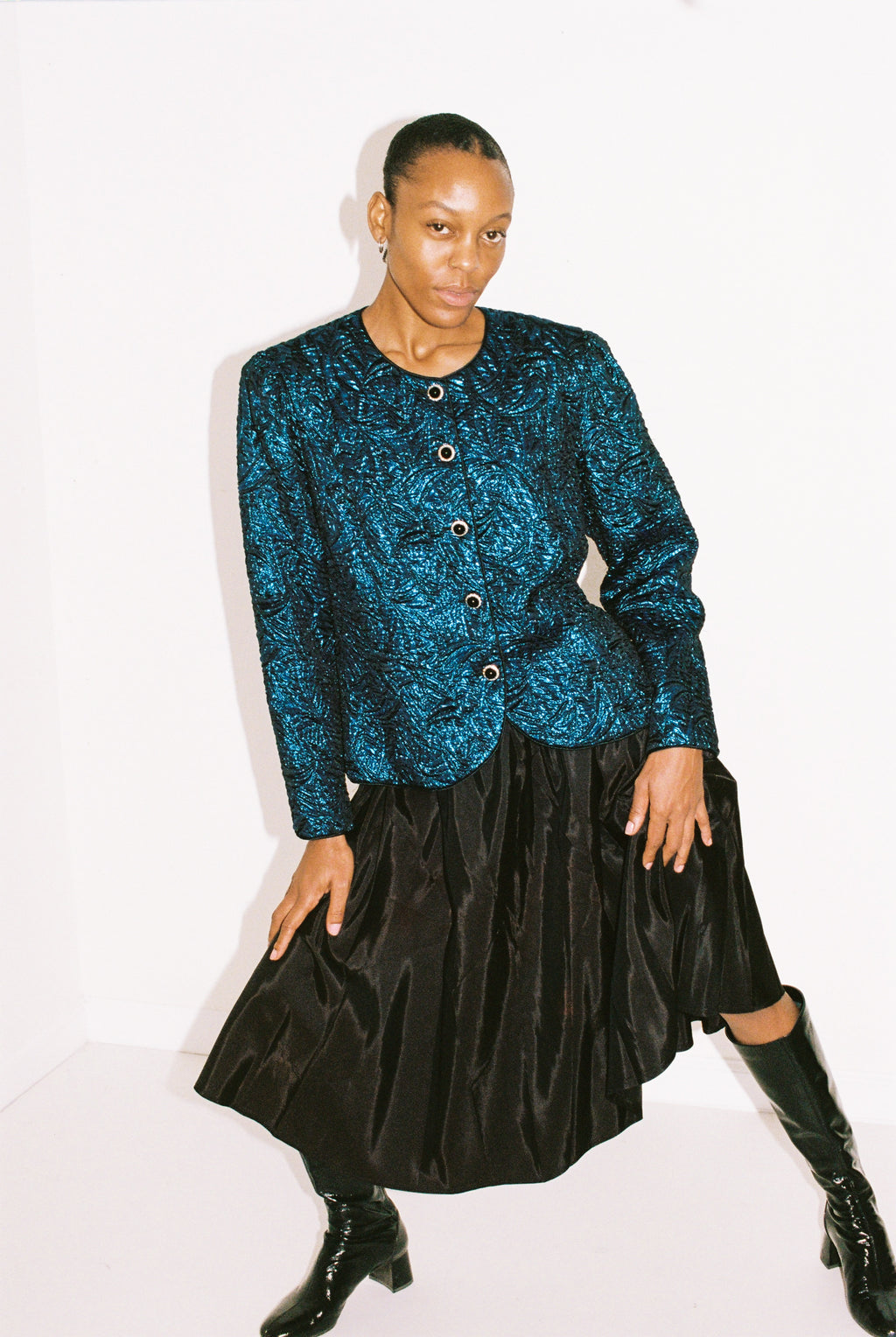 Person wearing a blue patterned top and black skirt with black boots on a white background