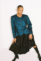 Person wearing a blue patterned top and black skirt with black boots on a white background