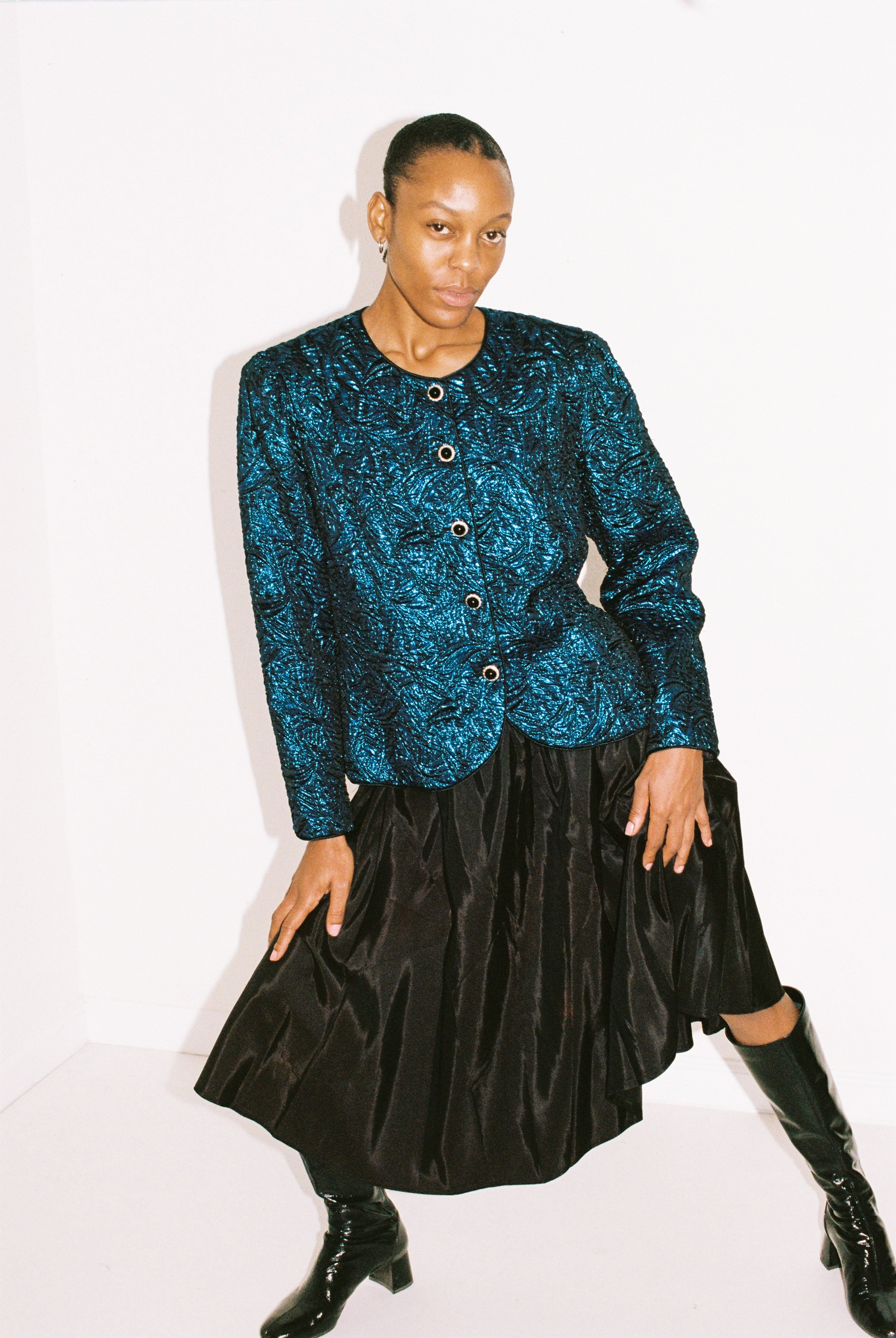 Person wearing a blue patterned top and black skirt with black boots on a white background