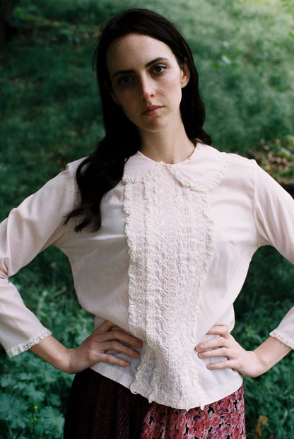 Woman wearing a light pink blouse with ruffled details against a green foliage background