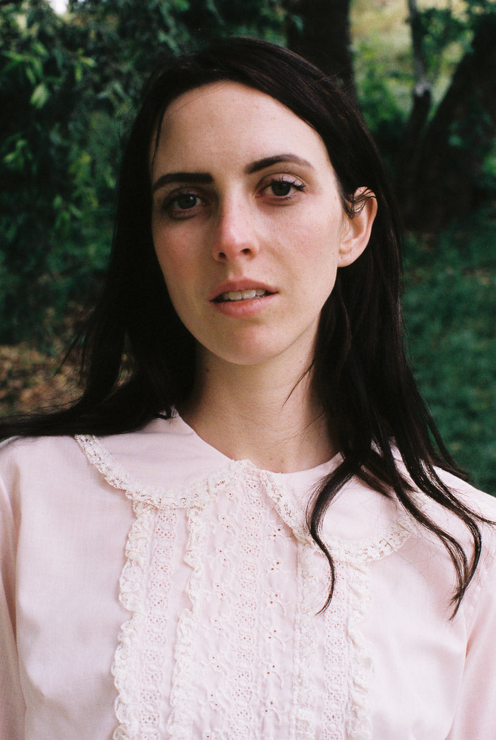 Woman with long dark hair wearing a light pink blouse against a blurred natural background