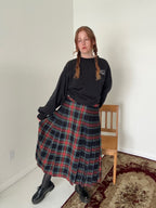 Person wearing a black sweater and plaid skirt standing in a room with a chair and carpet.