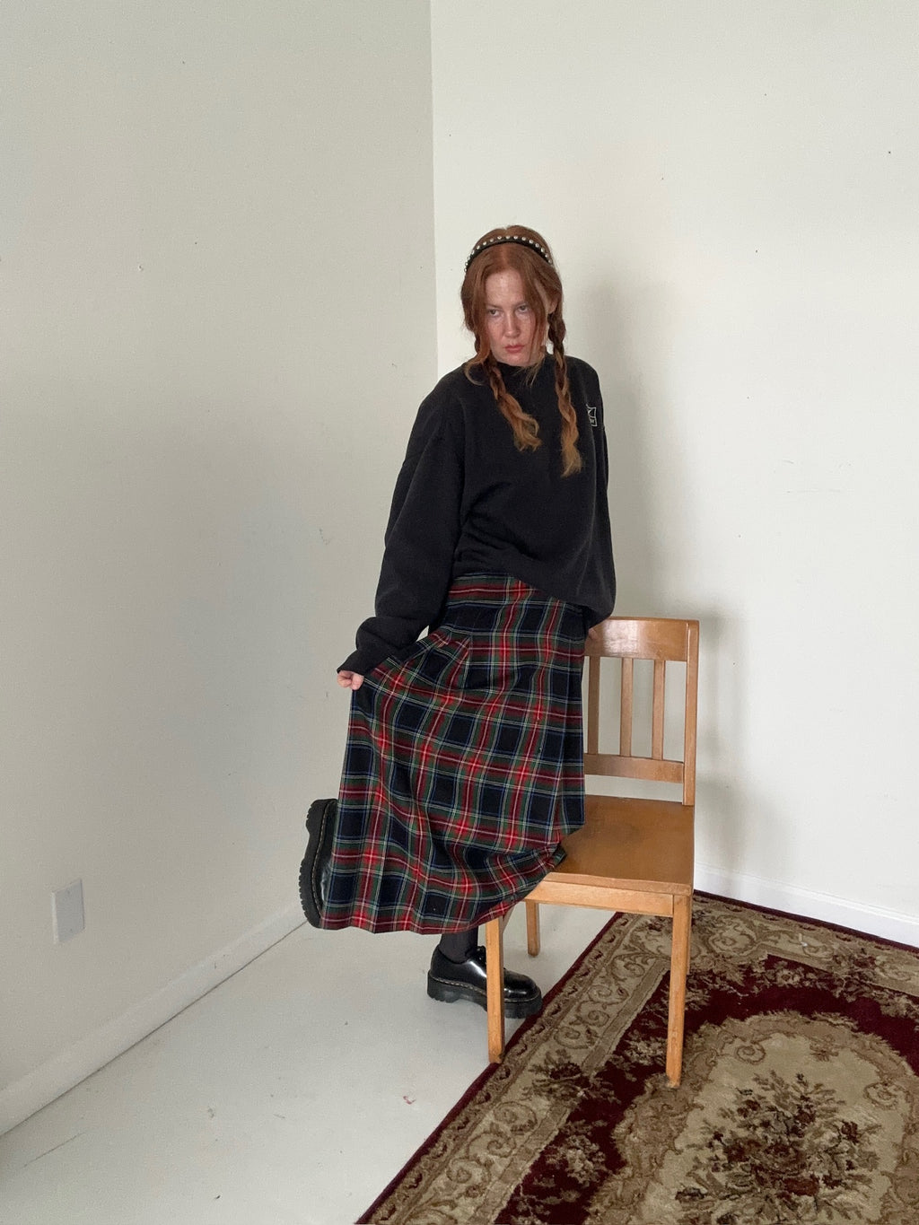 Person wearing a black sweater and plaid skirt standing next to a wooden chair against a white wall.