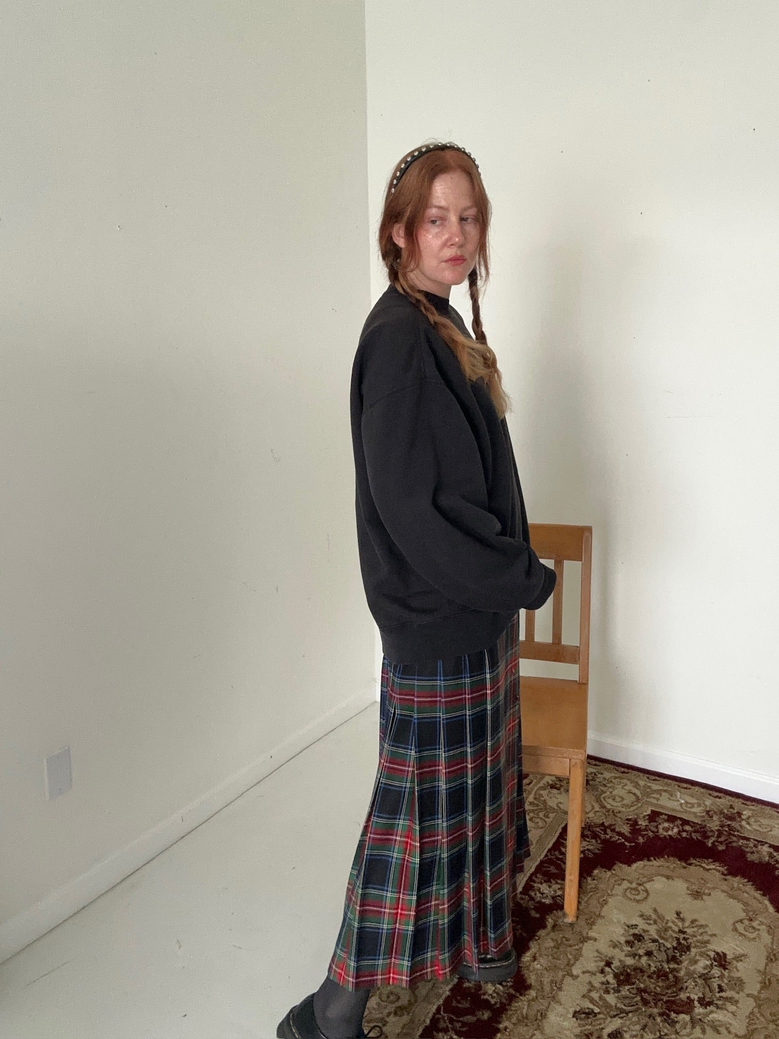 Person wearing a black sweater and plaid skirt standing in a room with a chair and carpet.