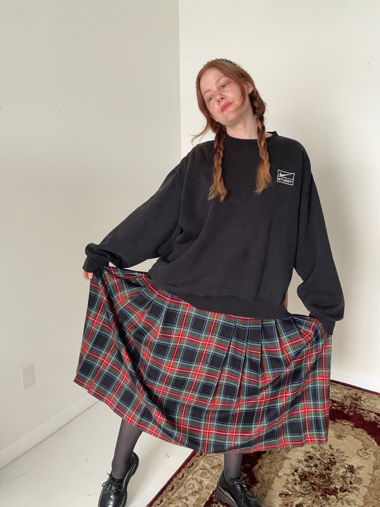 Person wearing a black sweatshirt and plaid skirt in a minimal indoor setting